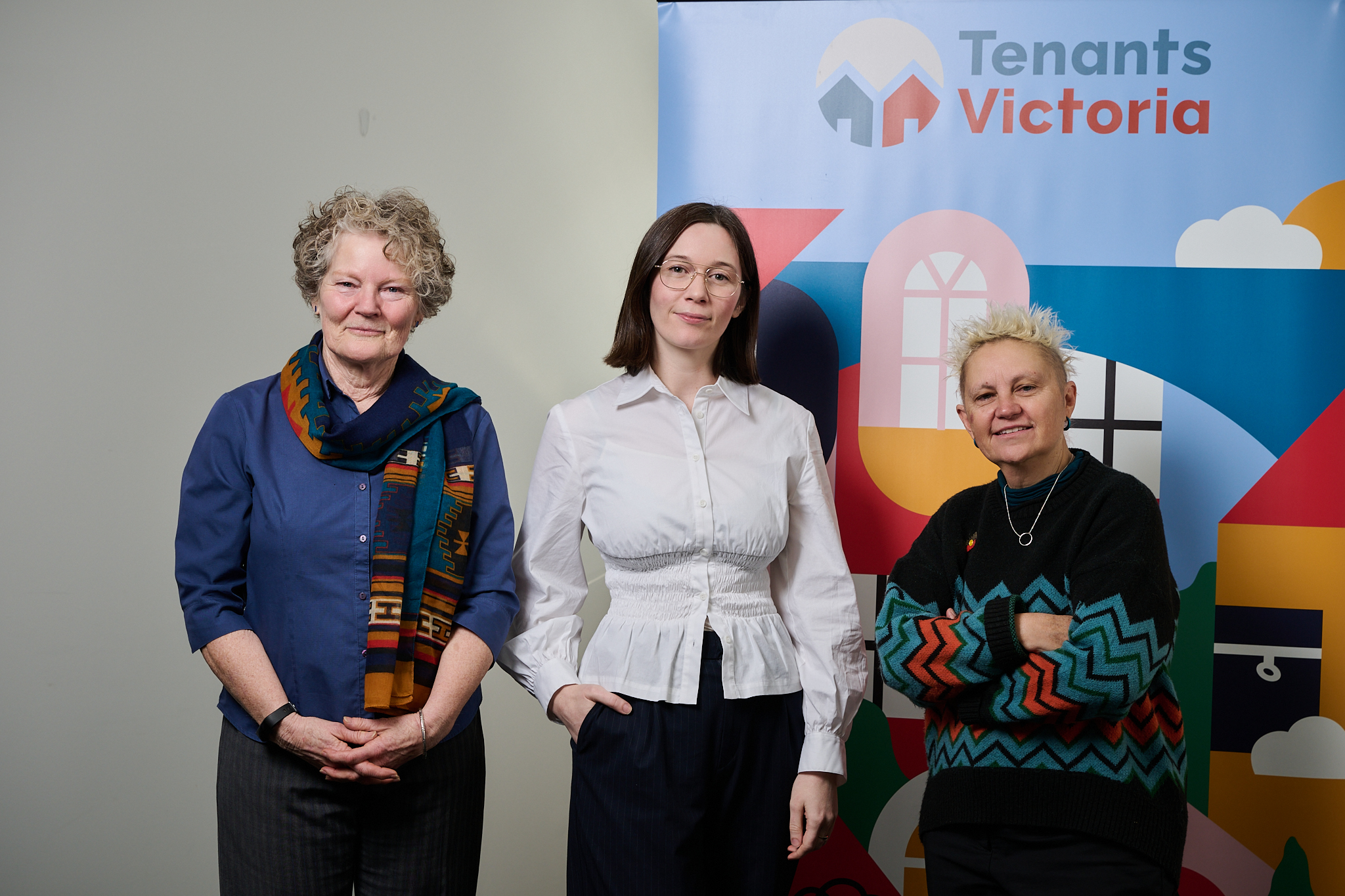 Three people standing in front of a 'Tenants Victoria' banner with colourful graphics of houses and clouds.