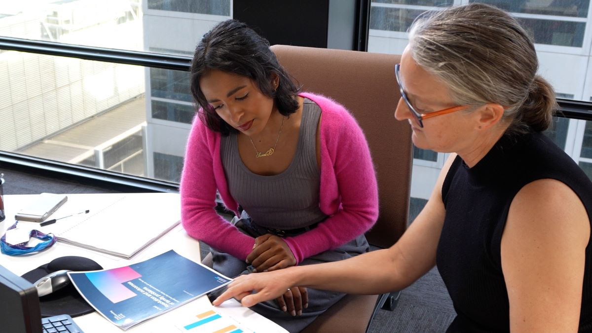 Two people in an office reviewing a document with charts and graphs at a desk with papers, a computer monitor, and glasses, with city buildings visible through large windows.