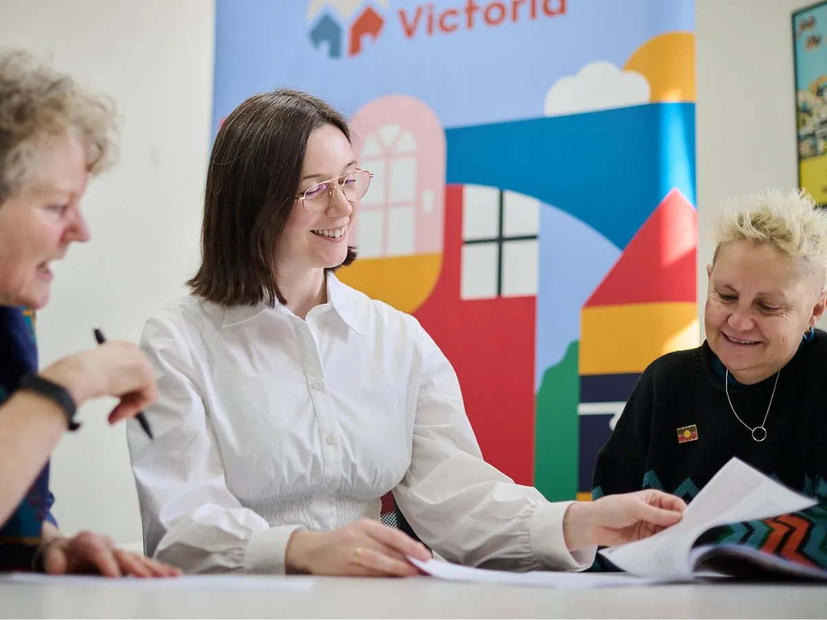 Three people sit at a table reviewing documents in a meeting setting. A colorful banner in the background displays stylized houses and rooftops. The person in the center wears a white shirt and holds a booklet, suggesting collaborative discussion.
