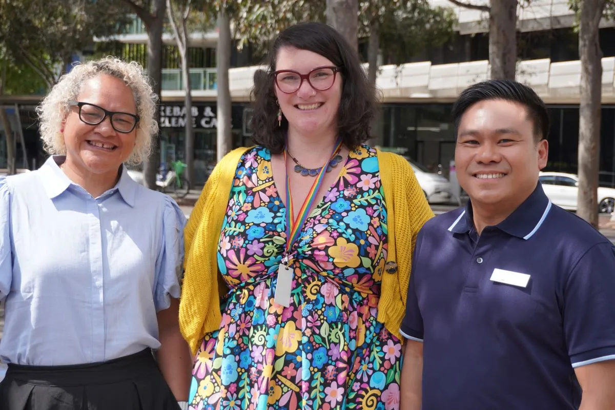 The image shows a group of people standing outdoors on a paved area near a modern building with glass panels. A few individuals are engaged in conversation, while others look towards the camera. Trees and greenery appear in the background.