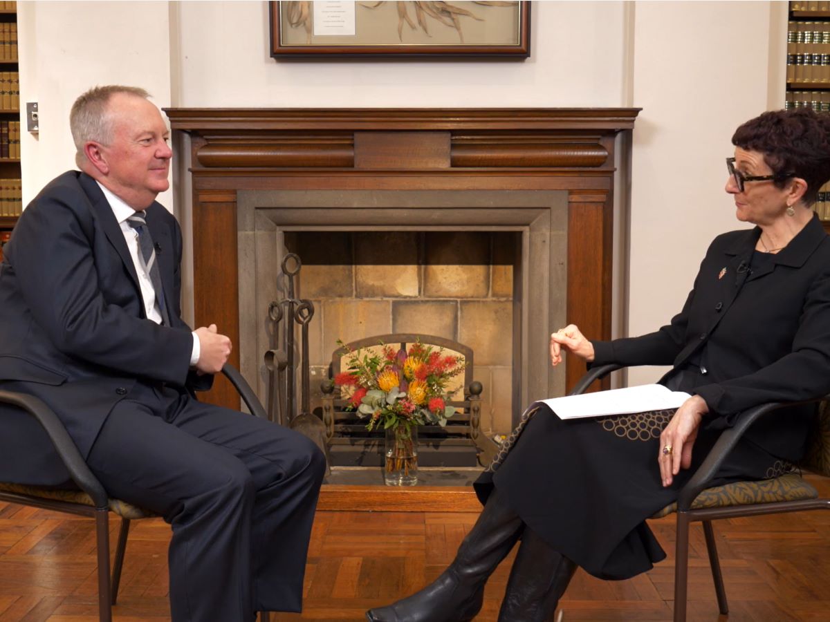 Two people sit facing each other in a formal room with wooden floors, bookshelves of legal volumes, and a fireplace. One person wears a suit and sits on the left, while the other, dressed in dark clothing, holds papers on the right. A flower arrangement sits before the fireplace area.Provide your feedback on BizChat