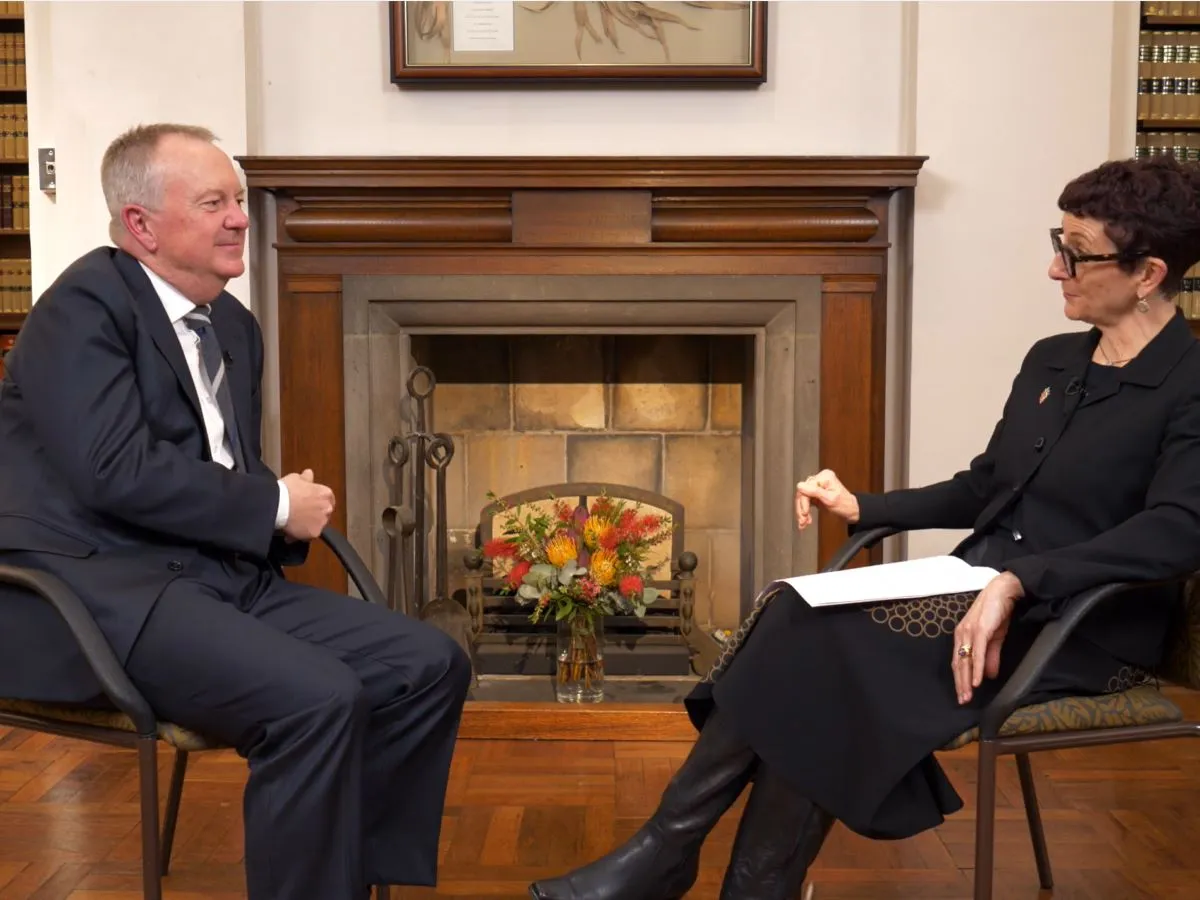 Two people sit facing each other in a formal room with wooden floors, bookshelves of legal volumes, and a fireplace. One person wears a suit and sits on the left, while the other, dressed in dark clothing, holds papers on the right. A flower arrangement sits before the fireplace area.Provide your feedback on BizChat