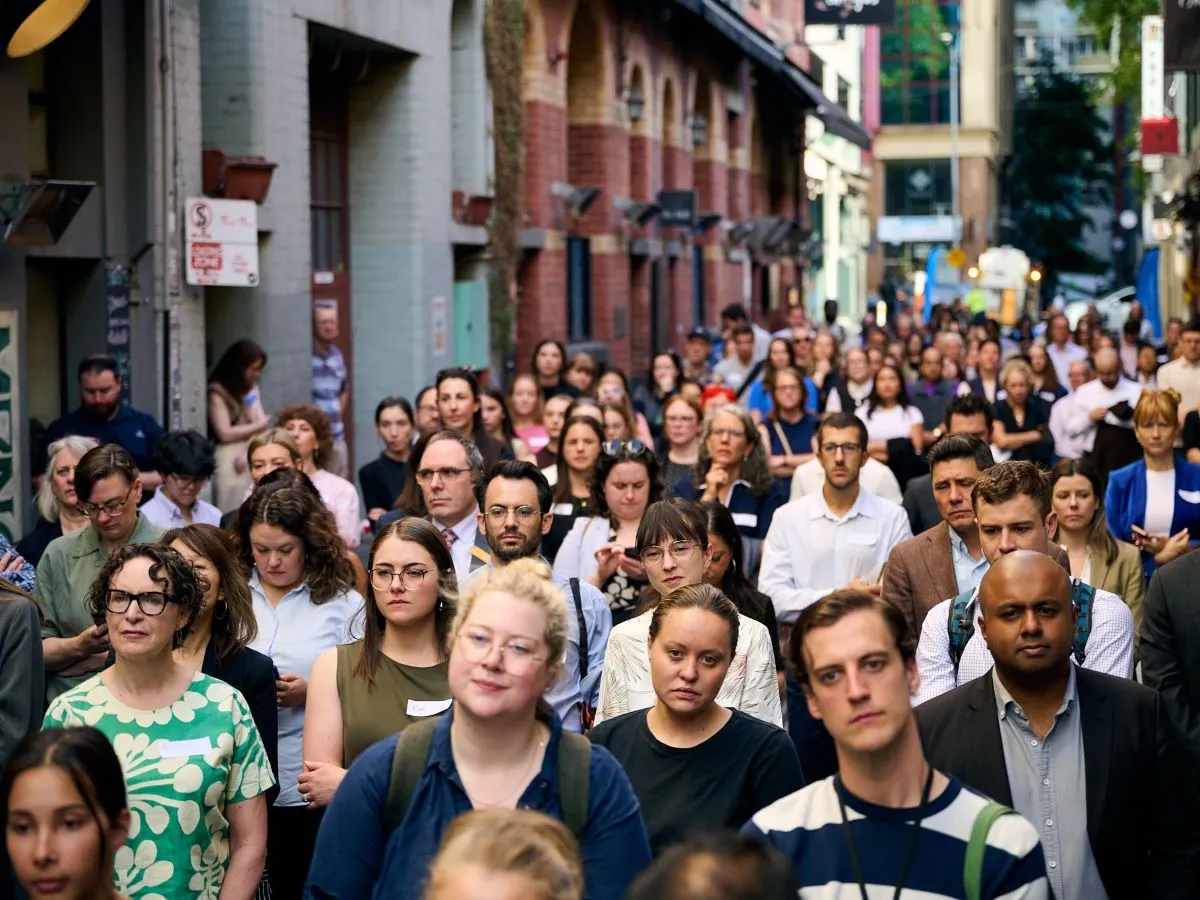 A large group of people gathered in a narrow city laneway for an outdoor event. At the front, a speaker stands at a podium next to a person providing sign language interpretation. Behind them is a blue Victoria Law Foundation banner. The crowd faces the stage, and the surrounding buildings create an urban backdrop with shopfronts and signage visible.