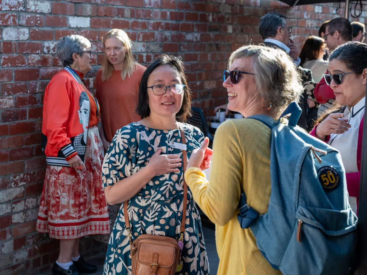 Several small groups speak in a courtyard with a red brick wall. People wear patterned, colourful clothing and carry bags or backpacks. A mix of seating and standing arrangements is visible. The background shows other attendees engaged in conversation.