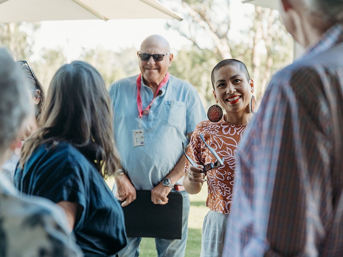 A small group stands outdoors under a sunshade, gathered in conversation. One person holds a tablet or clipboard, while another gestures with a hand raised and sunglasses in the other hand. Trees and bright daylight form the background. The group appears to be engaged in discussion in an open grassy area.