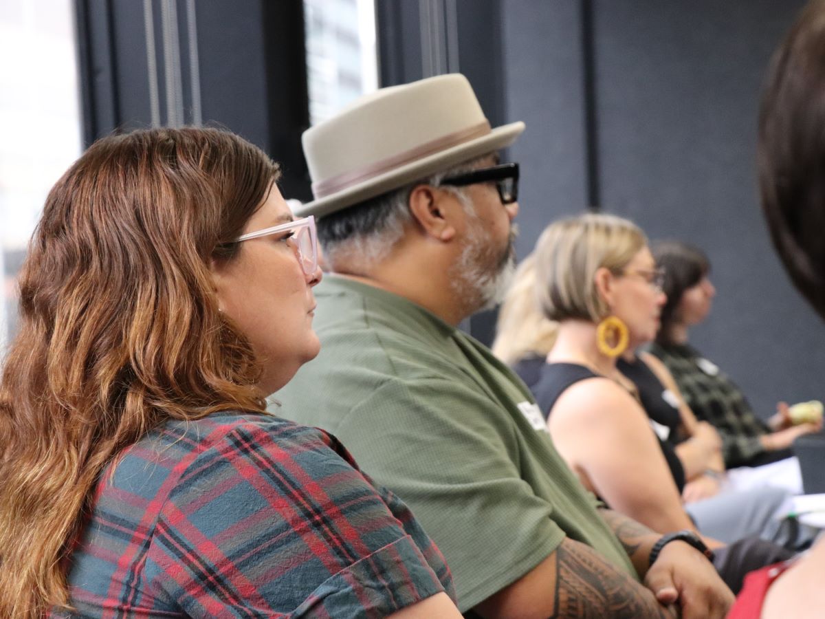 A group of people sit around tables in a meeting room with large windows and city buildings visible outside. Notebooks, cups, water jugs and laptops are on the tables. A presenter at the front gestures toward a projected slide showing text and graphics, while attendees listen and take notes.