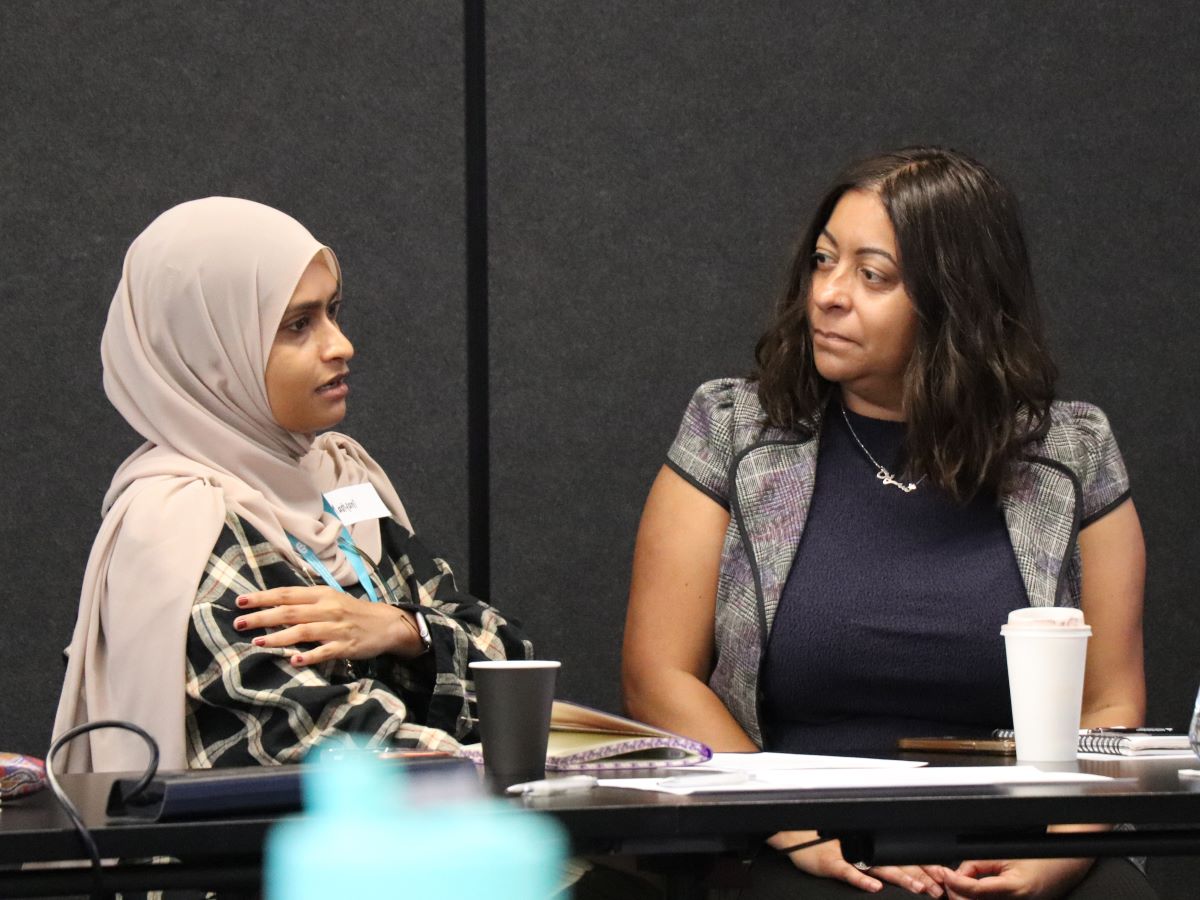 Two people sit at a meeting table in a conference room, engaged in conversation. One person wears a light head covering and patterned outer layer, while the other wears a short‑sleeved textured jacket. Notebooks, cups, and a takeaway coffee cup are on the table, suggesting an active workshop or discussion setting.