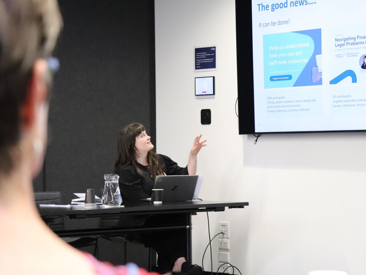 A person seated at a table presents to a group in a meeting room. The presenter gestures toward a projected slide displaying text and graphics. A laptop, cup, and water jug sit on the table. Another attendee is visible in the foreground, out of focus, watching the presentation.