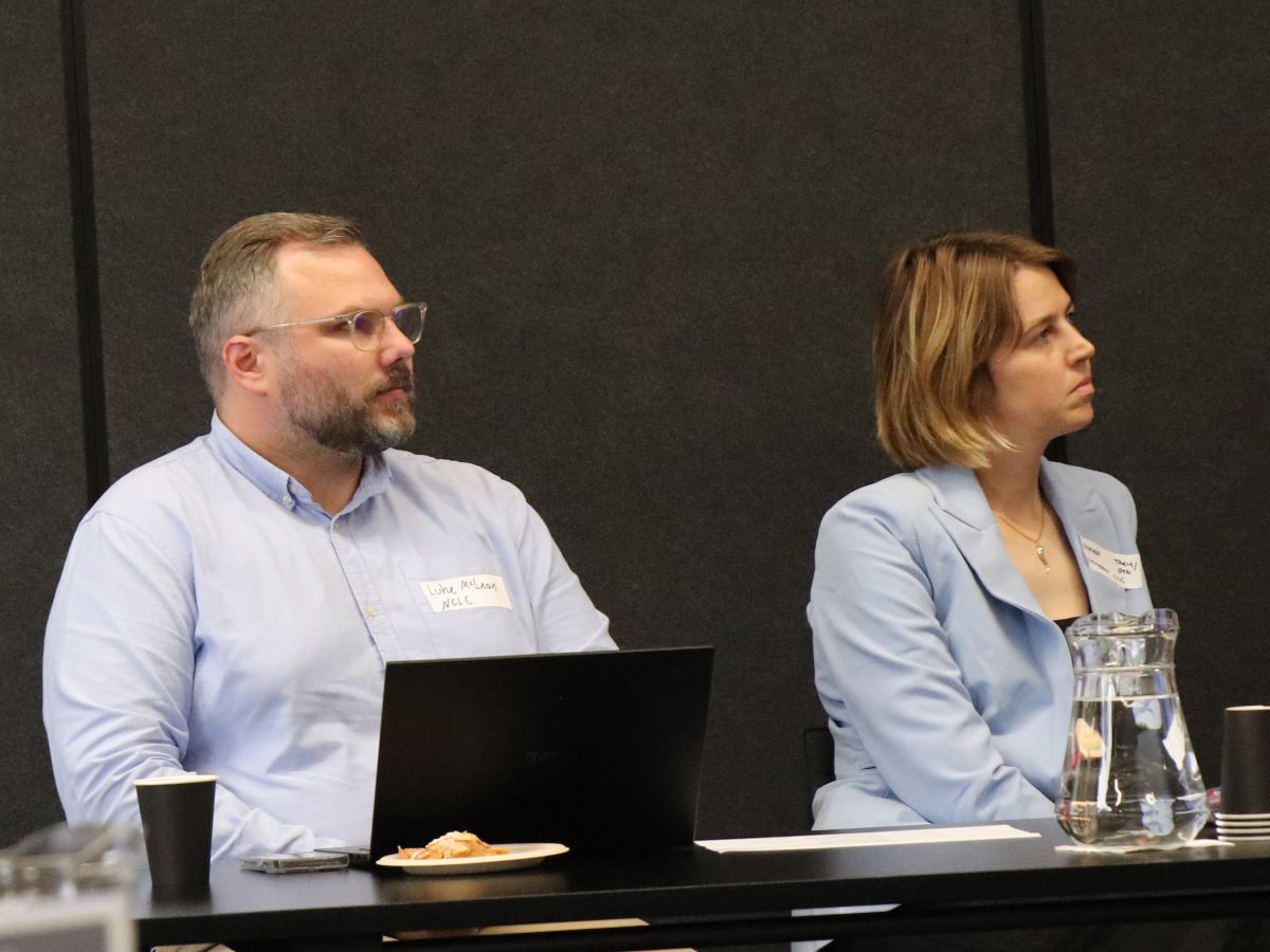 Two people sit at a meeting table in a conference room with dark walls. One person has a laptop open, with a cup, plate, phone and papers nearby. The other person sits beside a water jug and additional cups. Both appear to be listening during a formal discussion or workshop session.