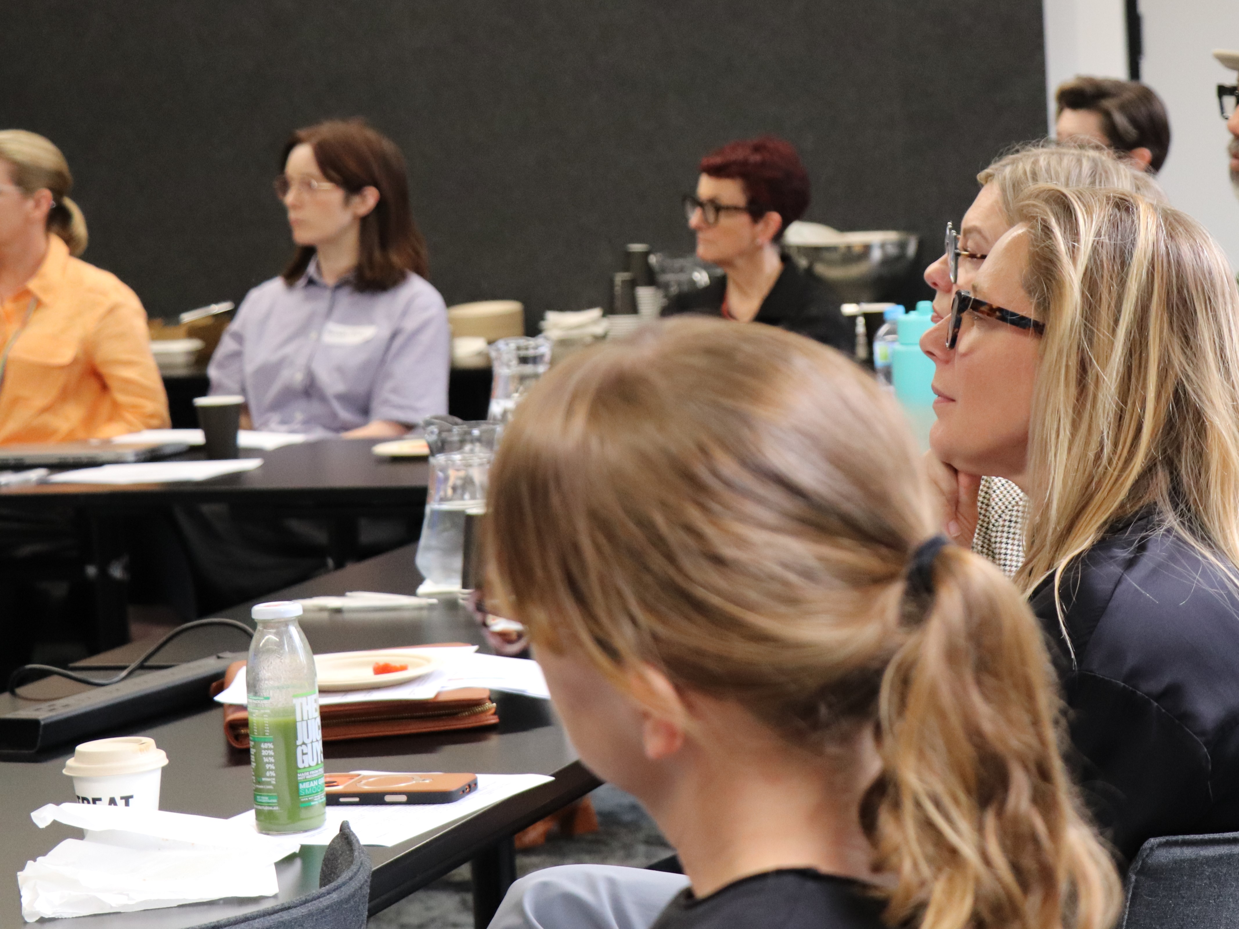 People sit around a large meeting table in a conference room, facing toward speakers or a presentation. Various drinks, notebooks, food containers and water jugs are on the tables. The setting appears organised and collaborative, with attendees seated in rows and groups, paying attention during a workshop or discussion sessio