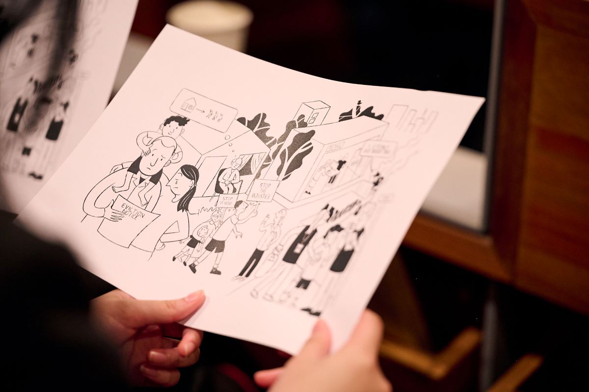 A person holds a sheet of paper showing a black‑and‑white illustration. The drawing features multiple cartoon-style figures interacting near buildings and trees. Some speech bubbles and signs contain simple shapes or unreadable text. The scene is viewed indoors, with blurred furniture and another similar sheet visible in the background.
