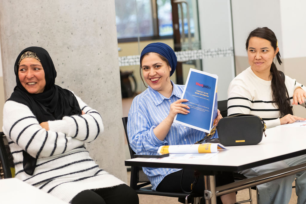 Three people seated at a table in a workshop setting; one holds up a blue booklet titled “Working Justice,” with notebooks, a pencil case, and papers on the table.