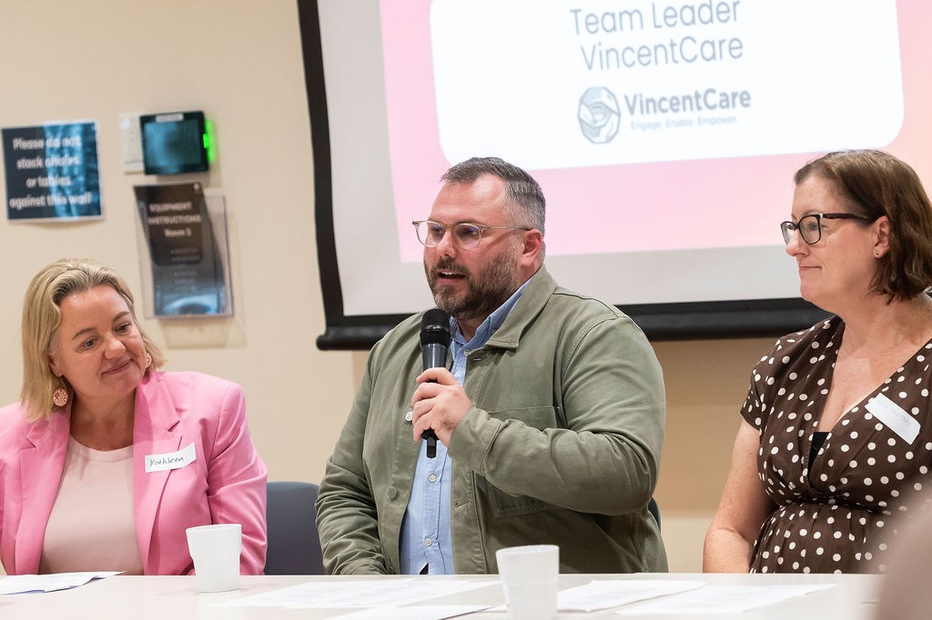 Panel discussion with three people seated at a table; one speaks into a microphone. Papers and cups on table; projection screen behind reads “Team Leader VincentCare.”