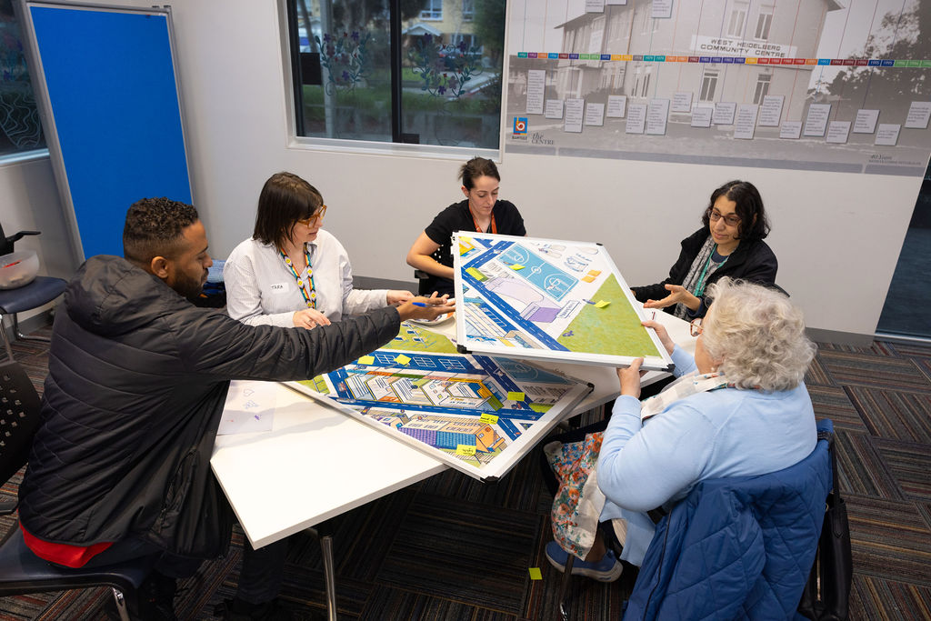 Five participants seated around a table reviewing large printed maps and plans; materials spread across table, with a window and wall chart in the background.