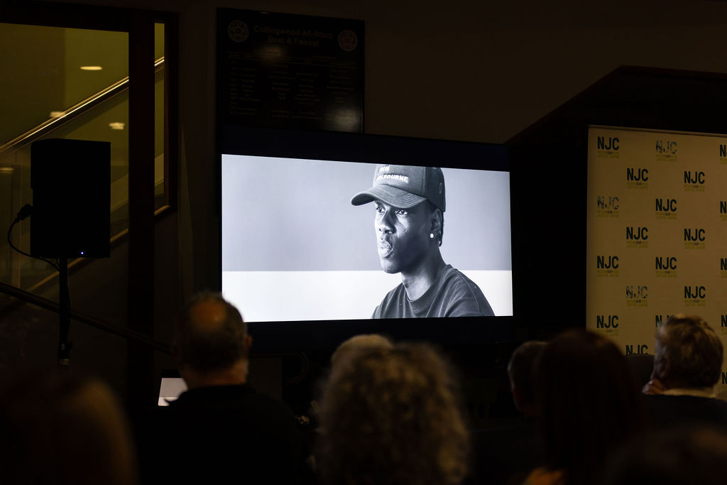 Audience seated in dim room watching a large screen showing a black-and-white portrait video; NJC signage and speaker setup visible near the screen.