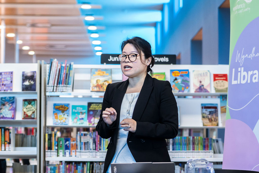 Person speaking in a library setting, standing near “Wyndham Libraries” banner; shelves of books and “Junior novels” sign visible in background.