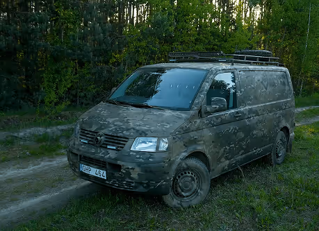 Camouflage-painted Volkswagen van parked on a grassy dirt path near a forest.