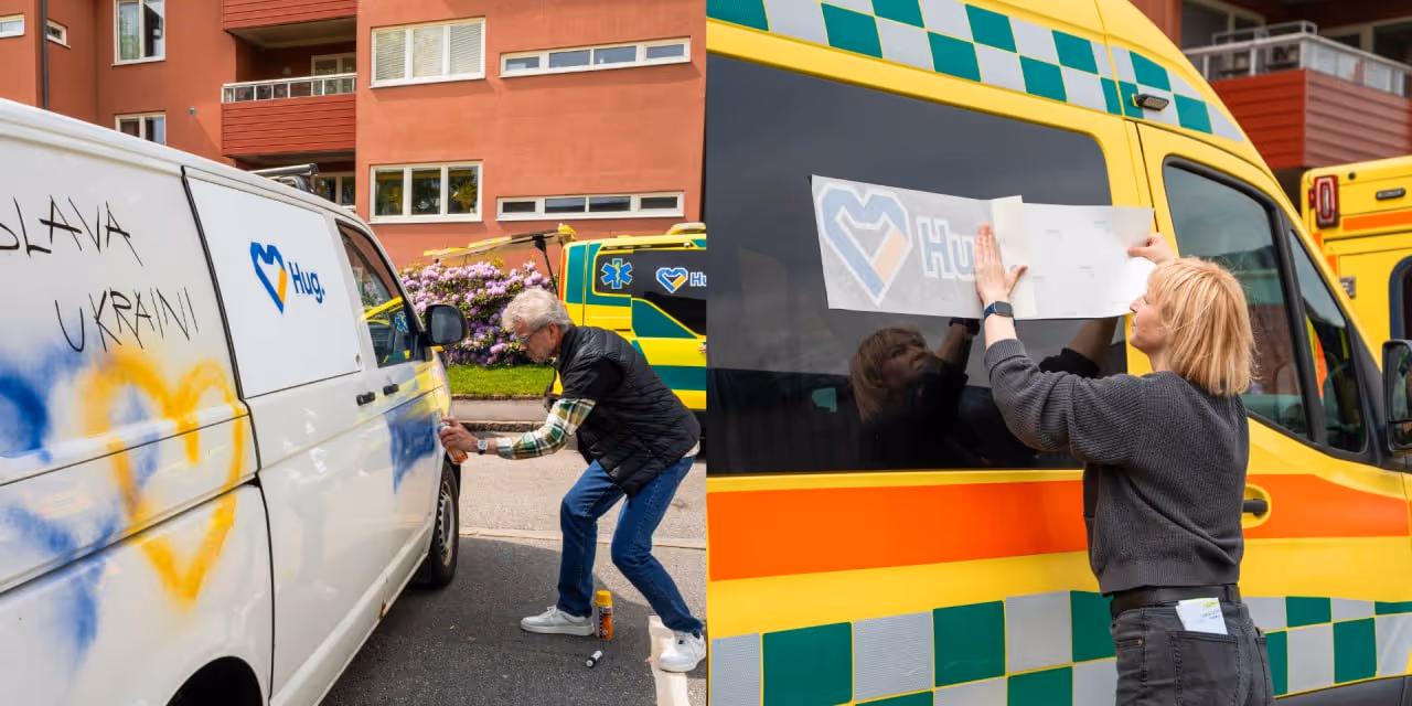 Two people applying a blue and yellow heart decal with the word 'Hug.' on the sides of white and yellow emergency vehicles.