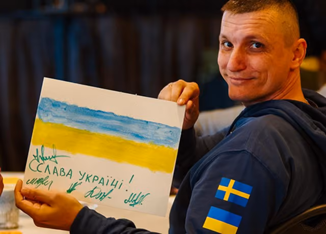 Man in a navy sweatshirt with Swedish and Ukrainian flag patches holding a painting of a blue and yellow flag with Ukrainian text.