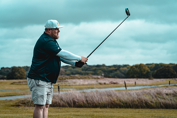 Man in a cap and shorts swinging a golf club on an outdoor golf course with cloudy sky.