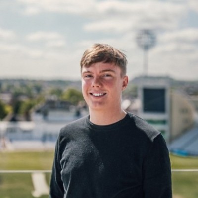 Smiling young man with short hair wearing a black shirt standing outdoors with a sports stadium in the background.