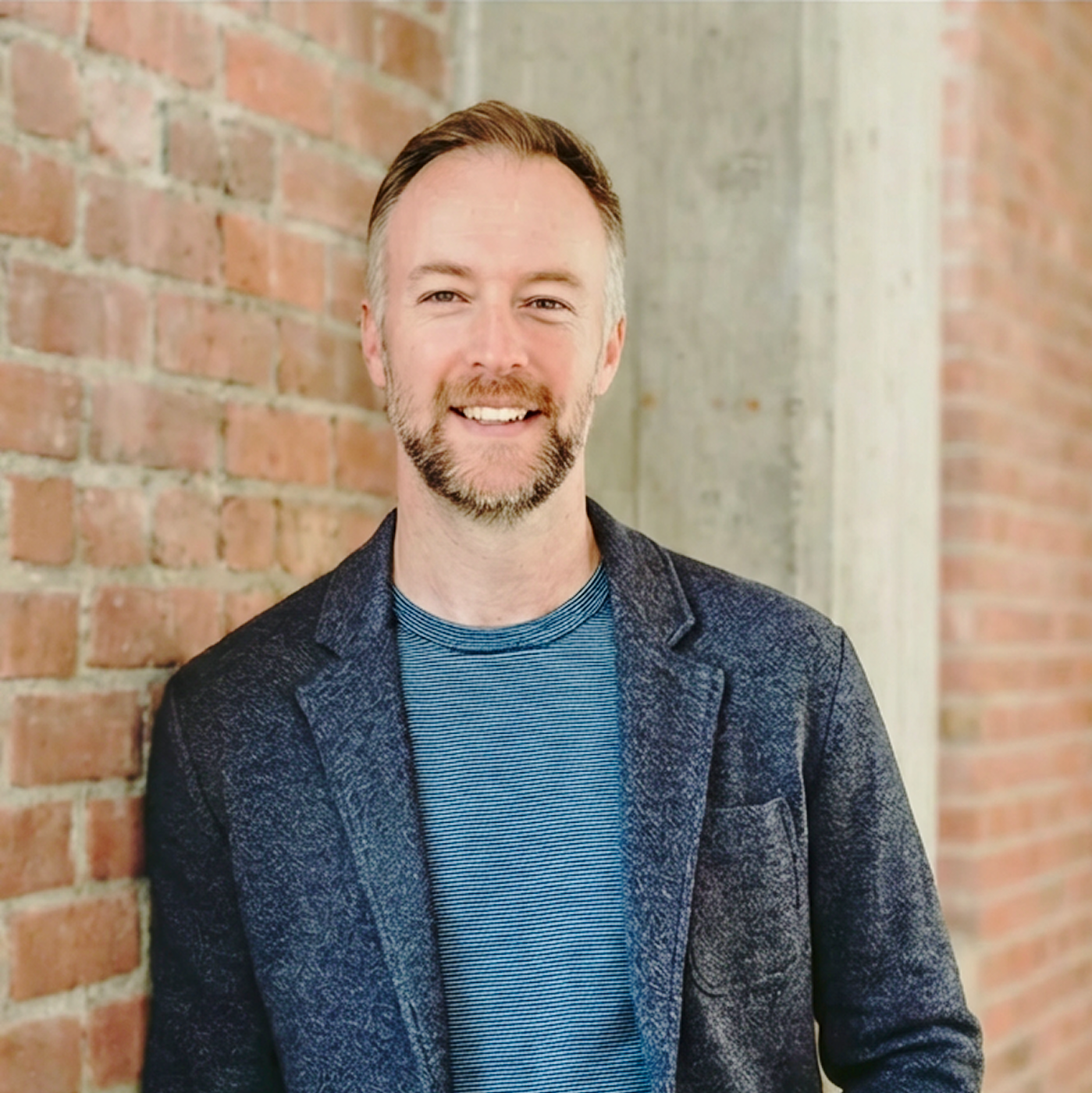 Smiling man with short brown hair and beard wearing a grey t-shirt and blue shirt, standing against a plain light background.