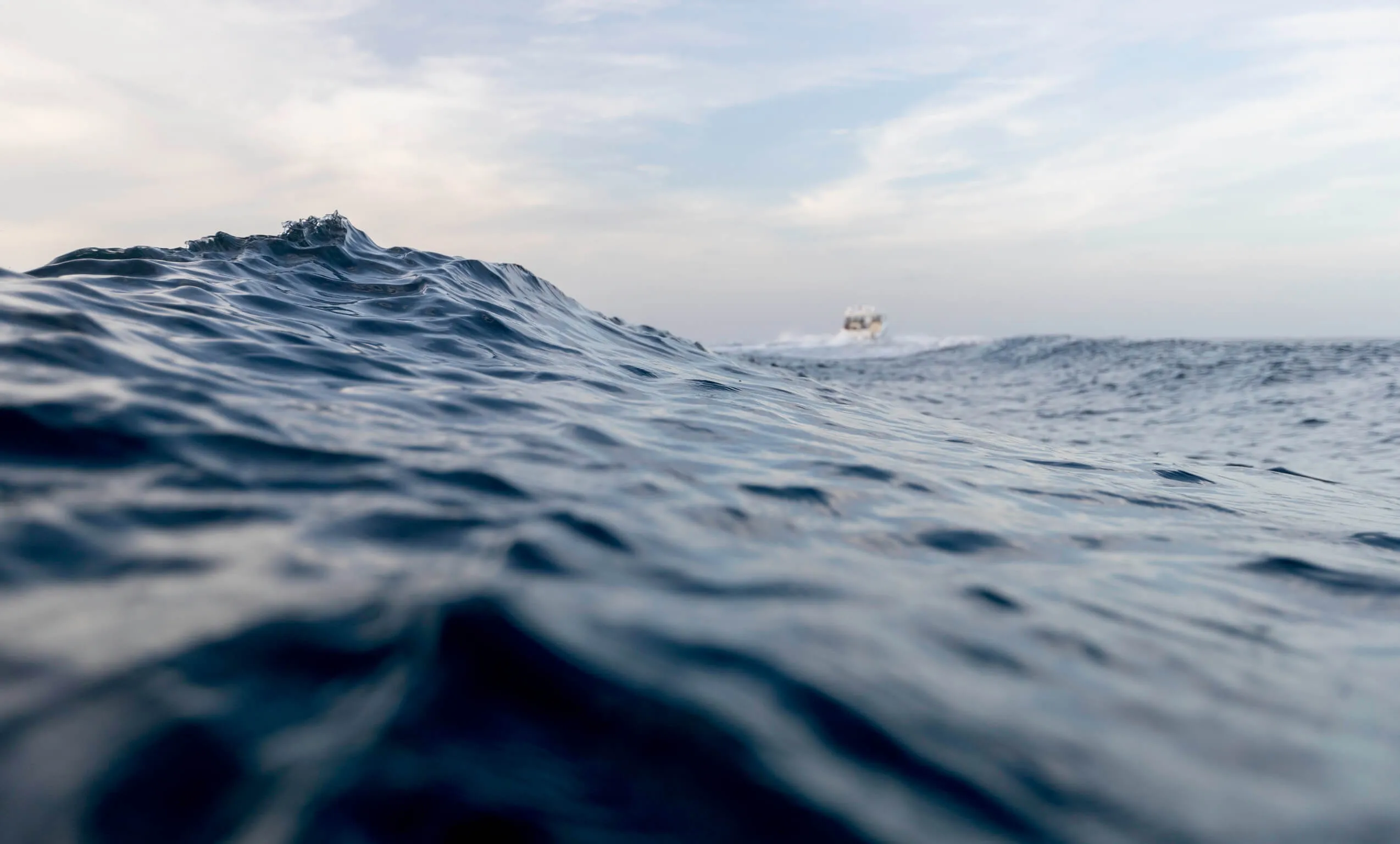 Close-up of ocean waves with a boat in the distant horizon under a partly cloudy sky.