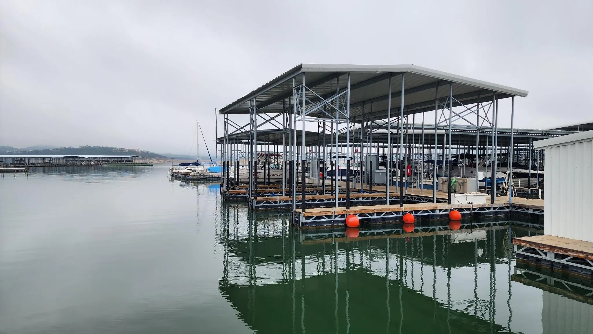 Covered boat docks with several boats moored on calm water under a cloudy sky.