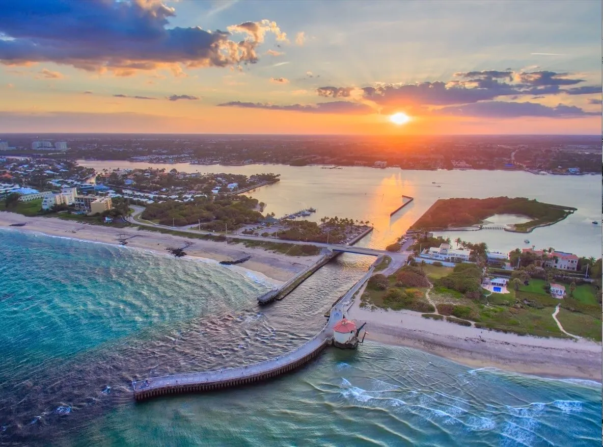 Aerial view of a coastal inlet at sunset with a pier, beach, bridge, and residential area.