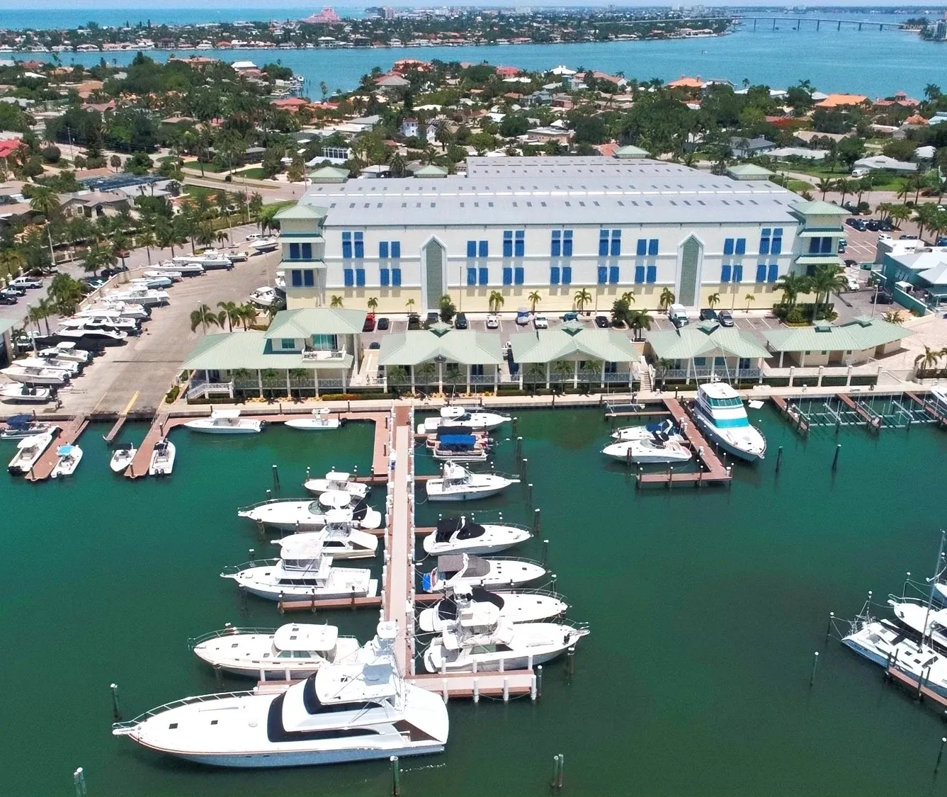 Aerial view of a marina with multiple white yachts docked, adjacent to a large building with greenish roofs and parking lot, with a residential area and water in the background.
