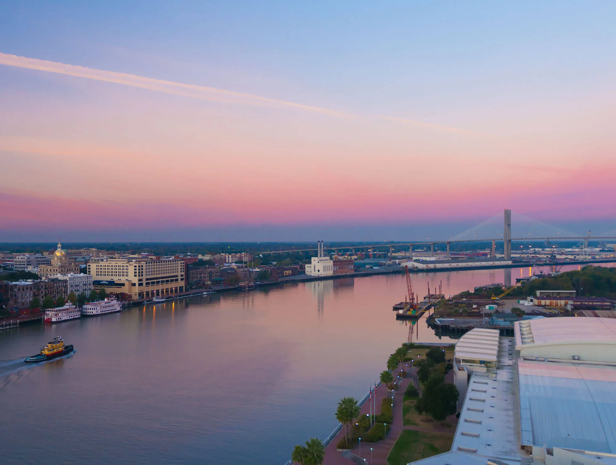 Aerial view of a riverfront city at sunset with a bridge spanning the river, boats on the water, and buildings lining the shore.