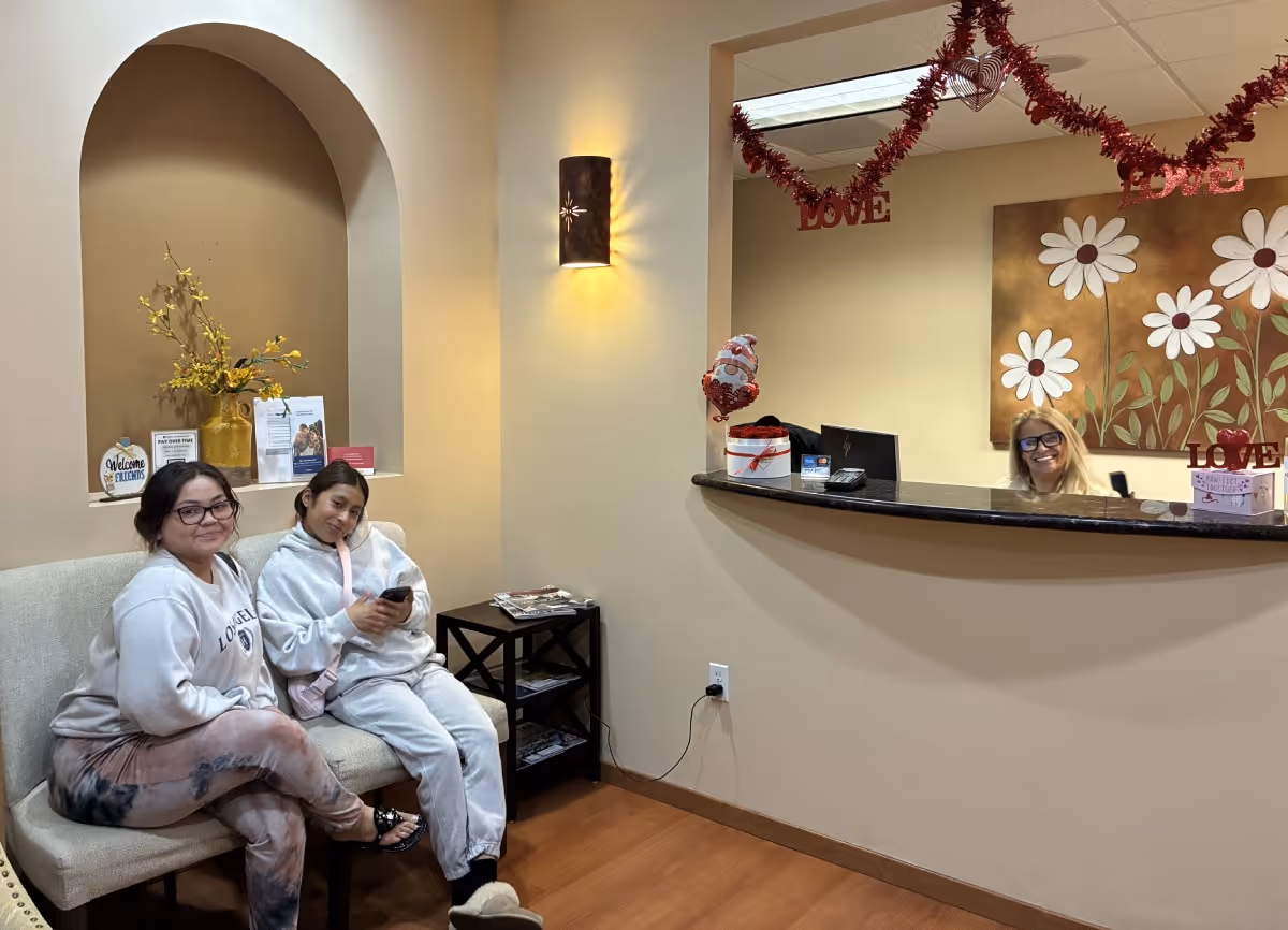 Two women seated on a beige bench in a warmly lit waiting area with a receptionist smiling behind a counter decorated with red garlands and 'LOVE' signs.