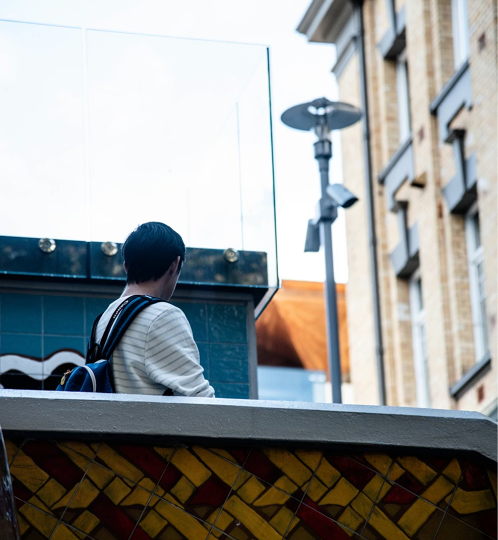 Personne avec un sac à dos debout à l'extérieur près d'un bâtiment avec un mur carrelé et une balustrade en verre.