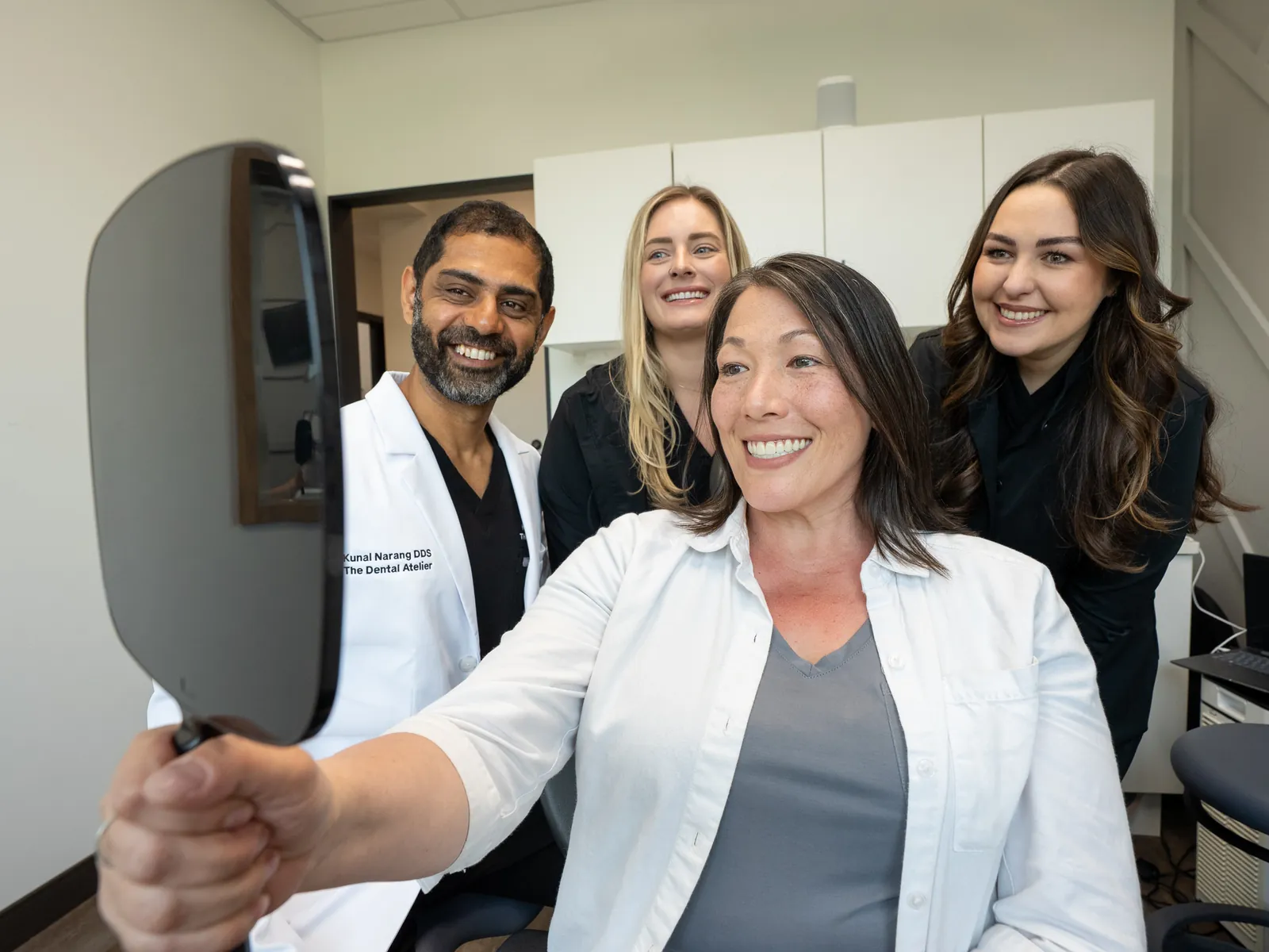 Smiling dental professional in black scrubs talking with a seated female patient in a clinic.