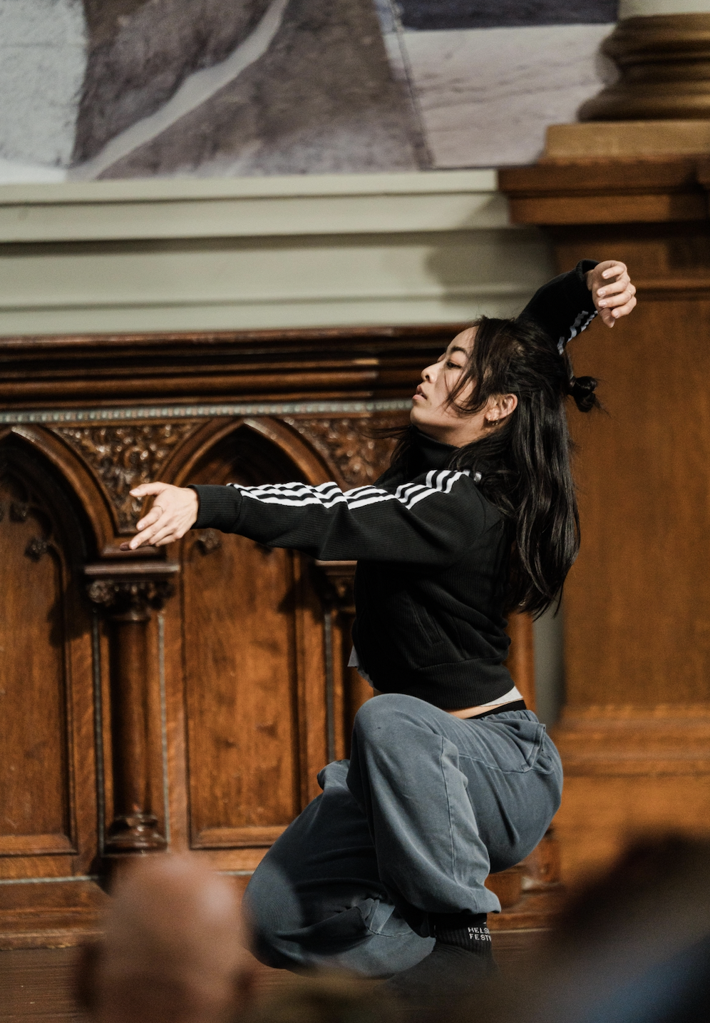 A young woman with long brown hair sits on the ground while resting her right elbow and left foot on a cement bench. She is looking off into the distance with a serene look.