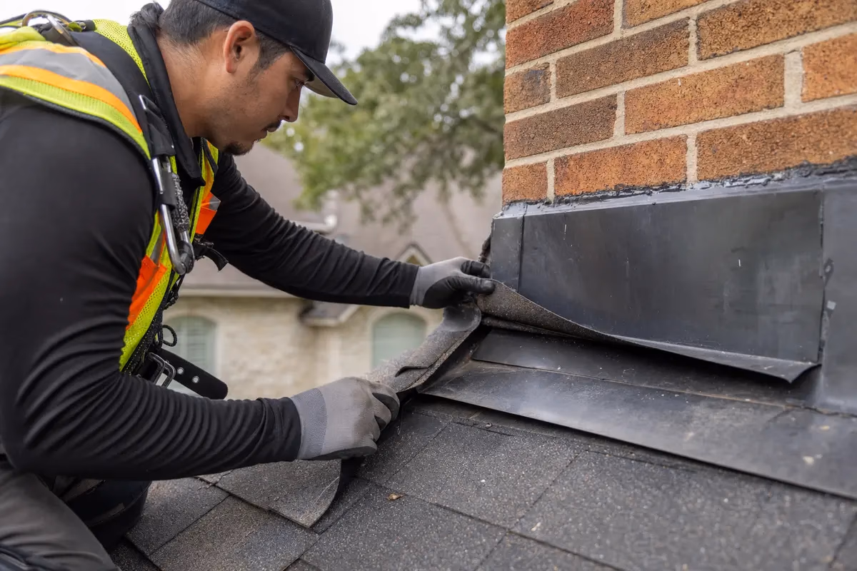 Close-up of roofer inspecting step flashing at a brick chimney with shingles lifted to show flashing seam