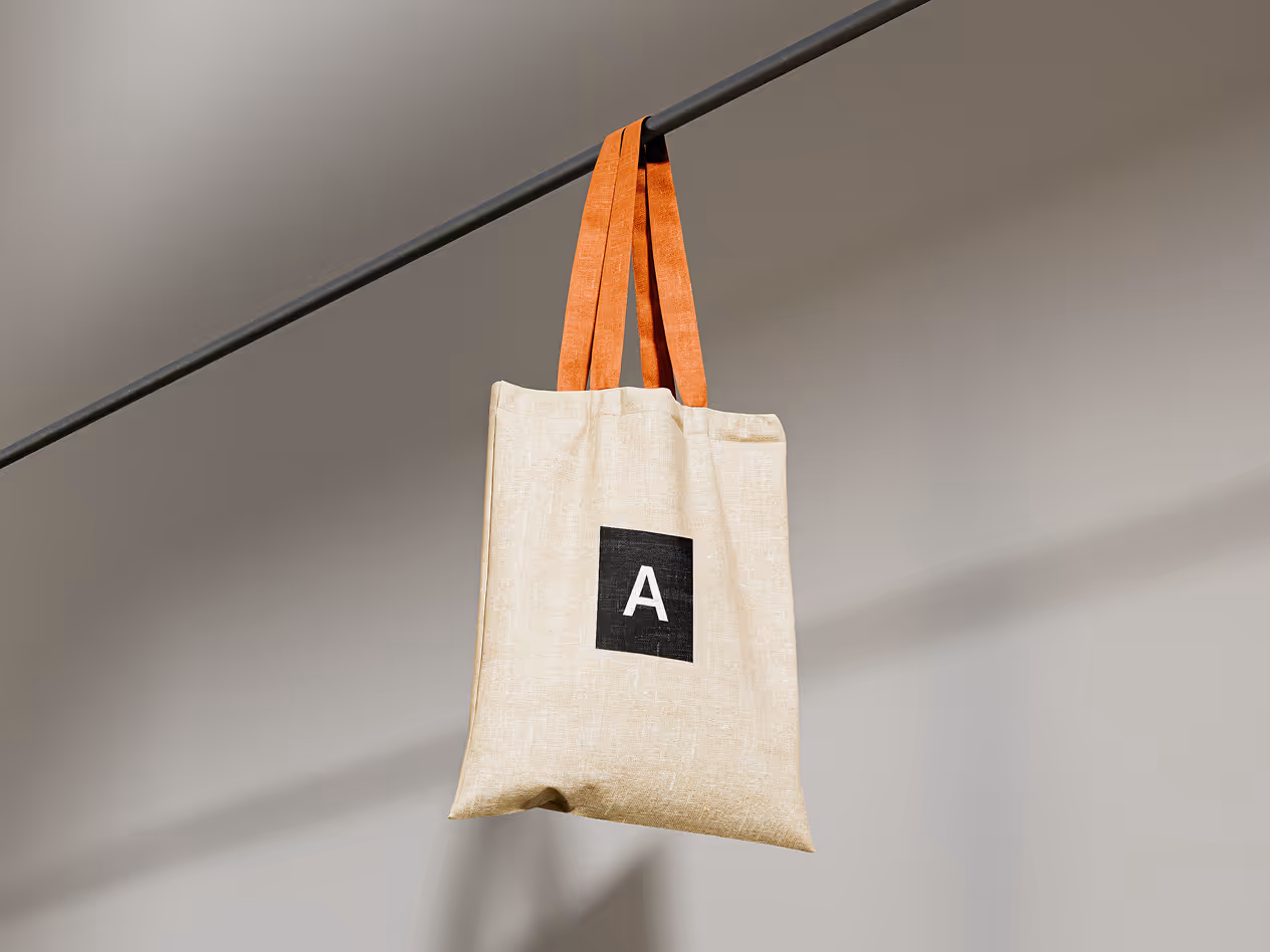 Beige fabric tote bag with orange handles hanging from a horizontal bar, featuring a black square logo with the letter “A” on the front.