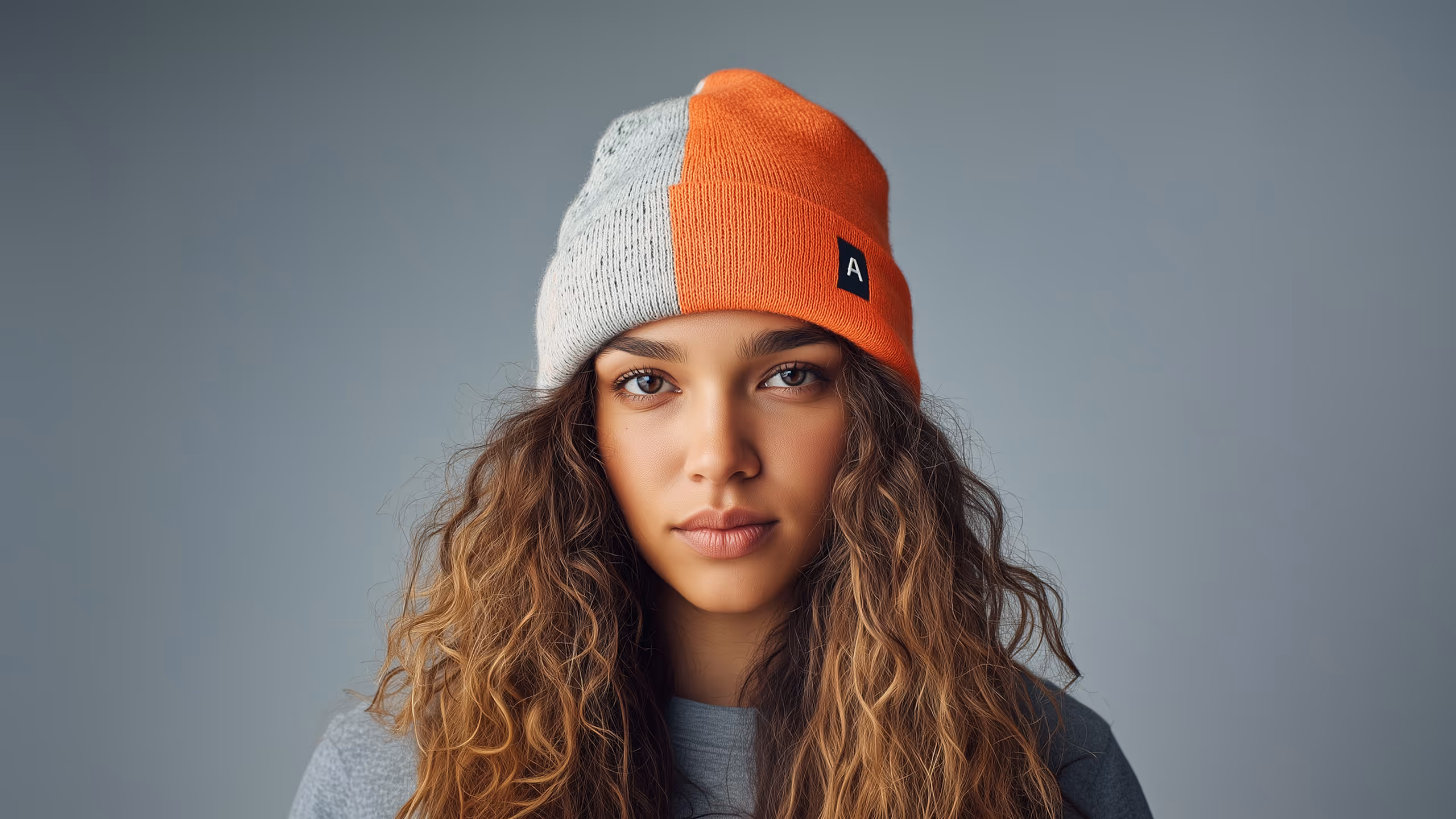 Portrait of a woman with curly hair wearing a two-tone gray and orange beanie against a neutral background.