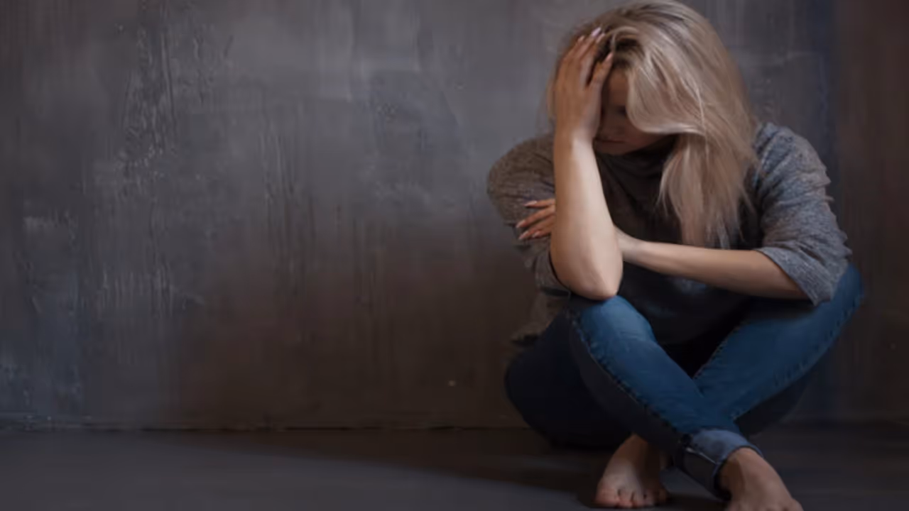 A woman in a dimly lit room, leaning against the wall alone, holding her head