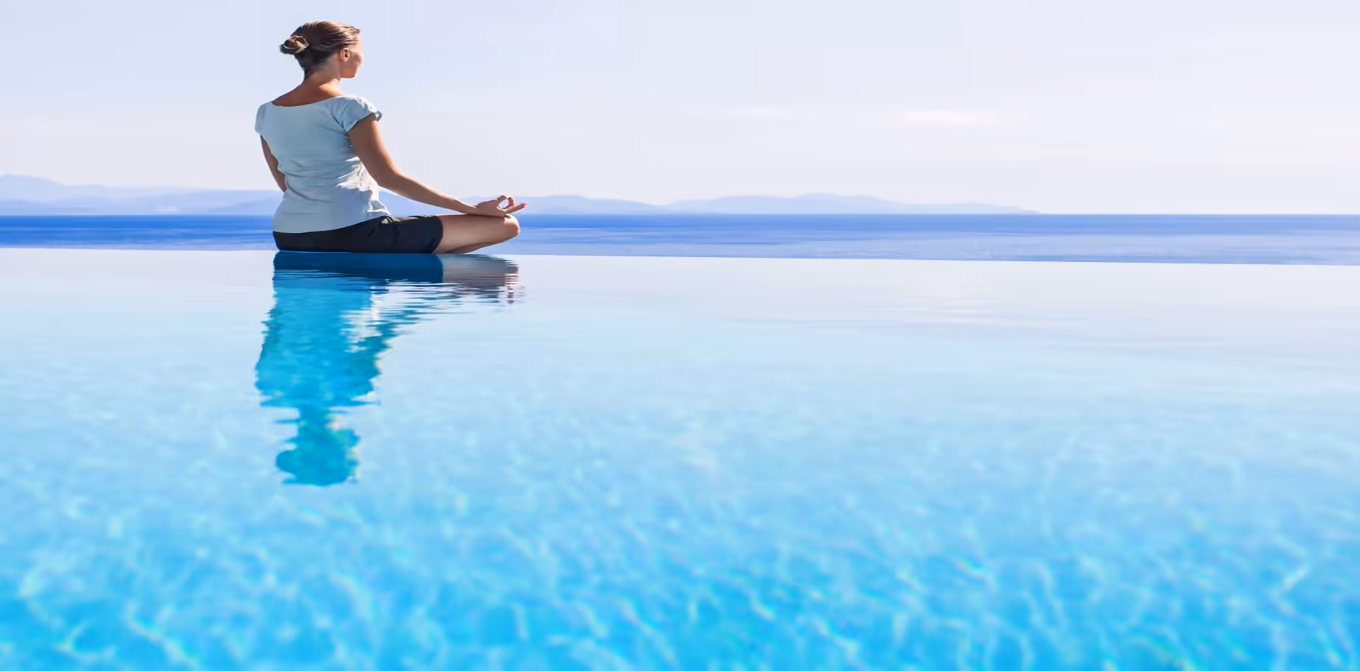 Yogi woman sitting on the edge of the pool facing the ocean in lotus position and meditating