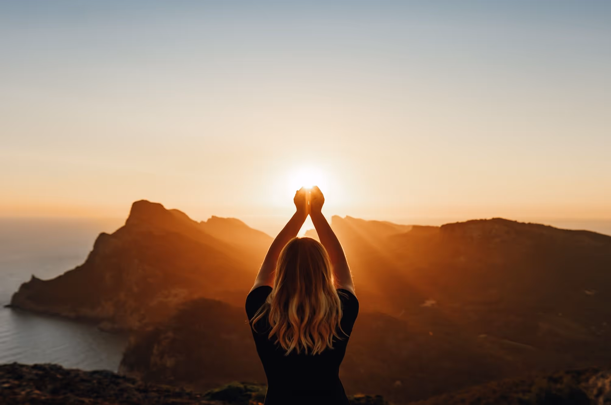A woman standing in front of the bay and the sea at sunrise, as if holding the sun in her hands.