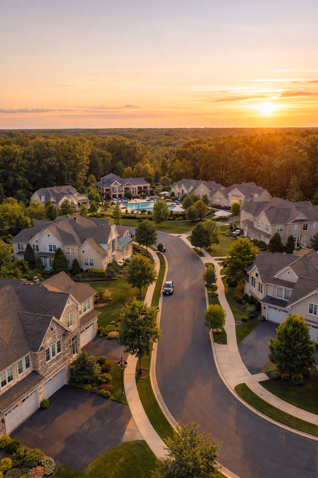 Sunset over suburban neighborhood with houses, trees, curved road, and a pool in the background.