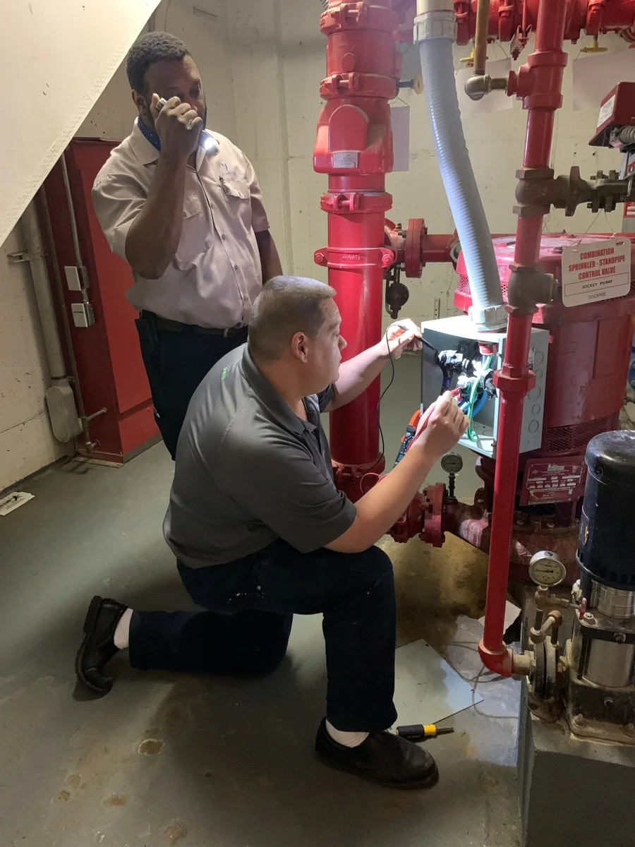 Two men inspecting and working on a red fire sprinkler standpipe system in a building's maintenance area, one kneeling and handling wiring inside a metal box, the other standing and holding a flashlight to illuminate the work area.
