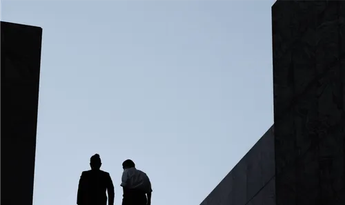 Silhouettes of two people standing between tall, dark buildings against a clear sky.