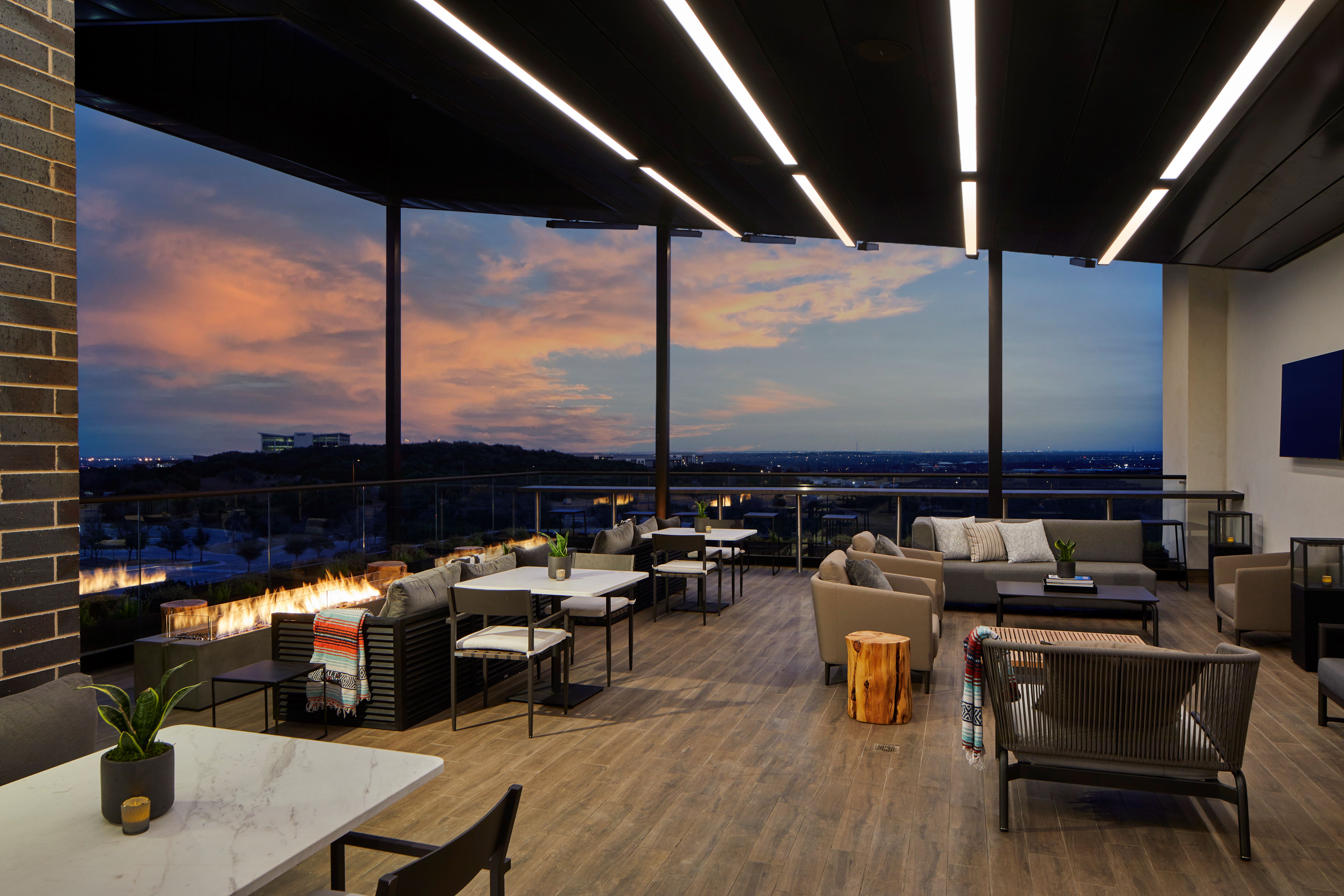 Modern outdoor rooftop lounge at dusk with wooden flooring, contemporary seating, fire feature, potted plants on tables, and a scenic view of distant hills and city lights under a colorful sky.