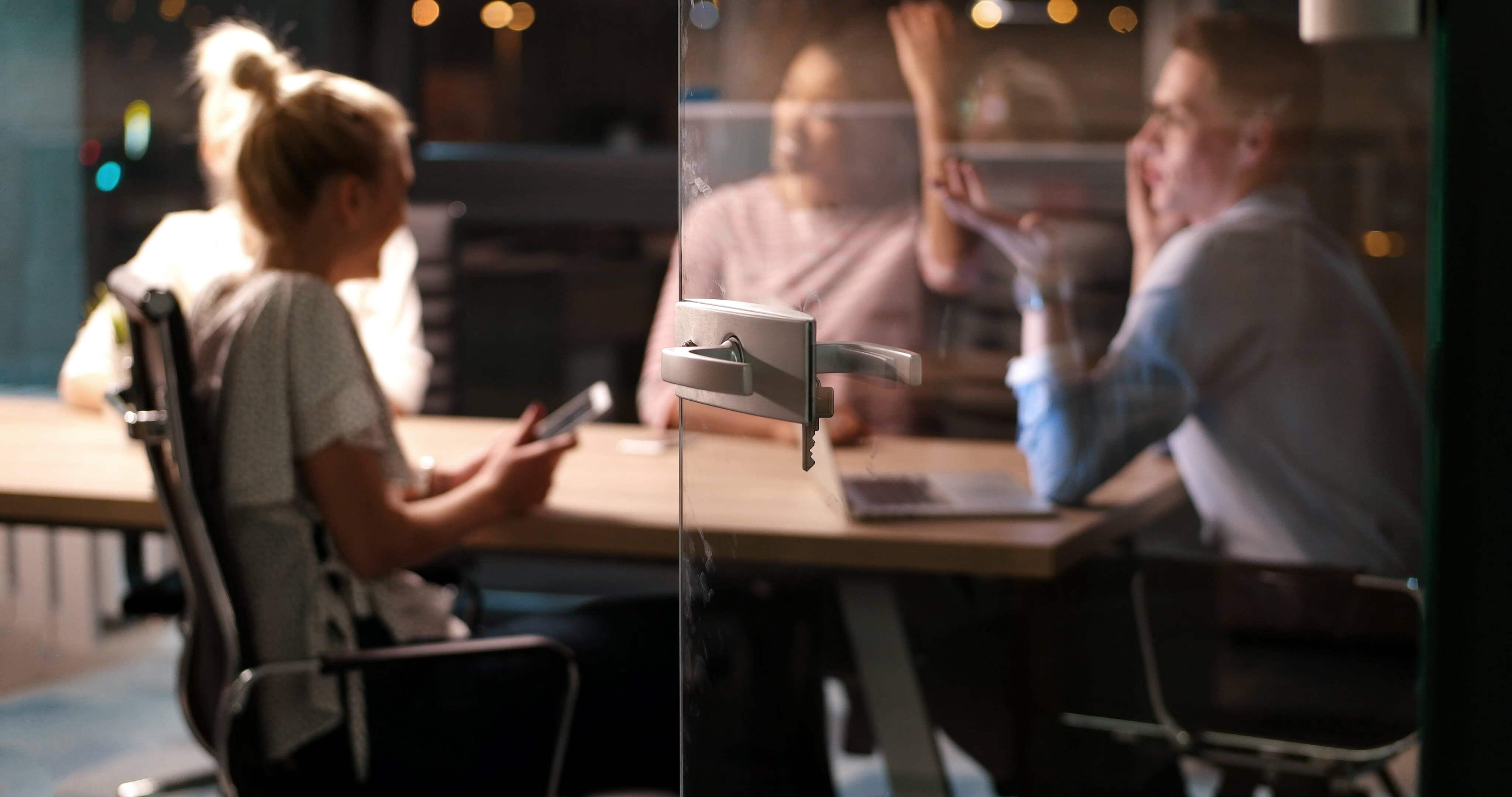 Three people sitting around a table engaged in a discussion, seen through a glass door with a metal handle and a key.