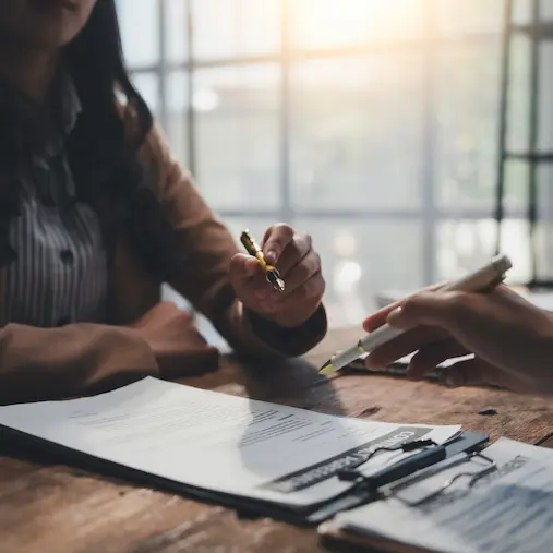 Two people discussing a document with pens over a wooden table near a bright window.