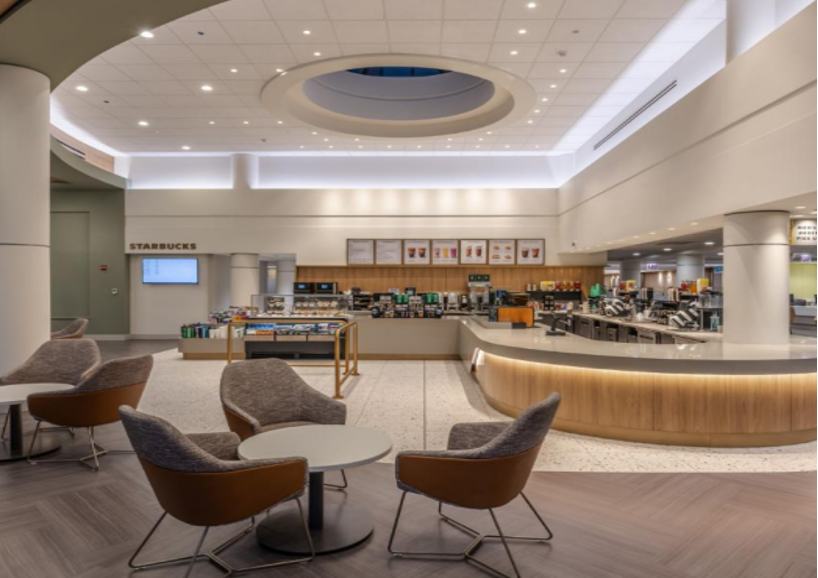 Modern Starbucks café interior with a curved service counter, light wood paneling, menu boards on the back wall, and several round tables surrounded by cushioned chairs in the seating area.
