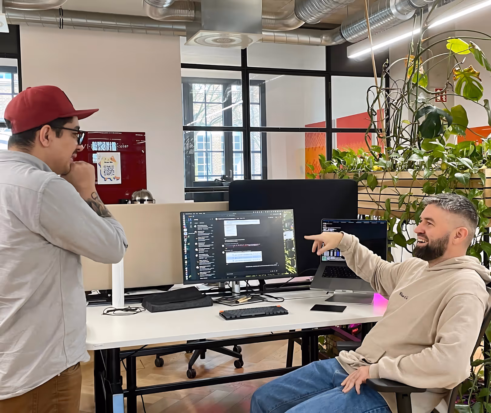 Two people discussing in front of a desk, while on the screen is code shown. Plants and office vibe in the background.