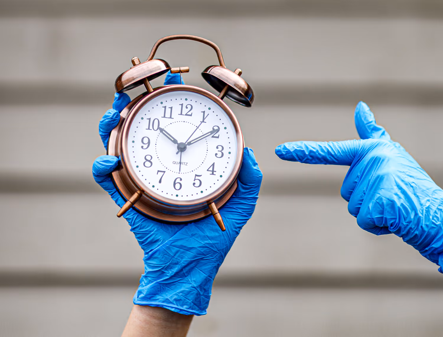 Hands with blue medical gloves on holding an alarm clock in one hand and pointing at the time on the other hand.
